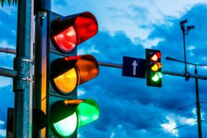 Lampu lalu lintas dengan tiga warna—merah, kuning, dan hijau—menyala bersamaan di sebuah persimpangan jalan saat senja, dengan latar langit biru dan kamera pengawas terpasang di tiang