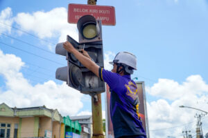 Petugas berseragam dan mengenakan helm sedang melakukan perbaikan atau pemasangan lampu lalu lintas di siang hari, dengan latar belakang langit biru dan bangunan toko.