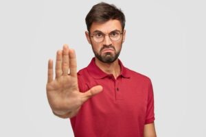 Pria berjenggot mengenakan kacamata dan kaos merah dengan ekspresi serius mengangkat tangan ke depan seolah memberi isyarat stop sebagai simbol peringatan terhadap pelanggaran