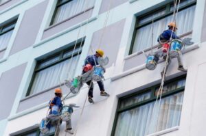 Tiga pekerja sedang mengecat dinding luar gedung bertingkat menggunakan tali dan kursi gantung.