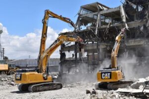 Dua alat berat excavator merek CAT sedang melakukan pembongkaran bangunan bertingkat yang sudah rusak parah, dengan puing-puing bertebaran di sekitar lokasi di bawah langit cerah