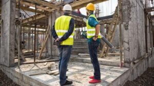 Dua pria memakai rompi reflektif dan helm keselamatan sedang berdiskusi di area proyek konstruksi bertingkat yang masih dalam tahap struktur beton.