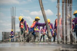Sejumlah pekerja konstruksi mengenakan helm kuning, rompi keselamatan, dan pakaian pelindung sedang menuangkan beton di area proyek pembangunan dengan langit cerah di latar belakang
